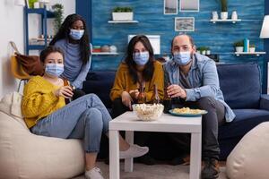 Pov of african woman and friends looking at tv holding remote control wearing face mask because of coronavirus pandemic outbreak drinking beer, relaxing, sitting on sofa in living room. photo