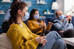 Mixed races of friends looking at tv, man holding remote control wearing face mask because of coronavirus pandemic outbreak drinking beer, relaxing, sitting on sofa in living room. photo