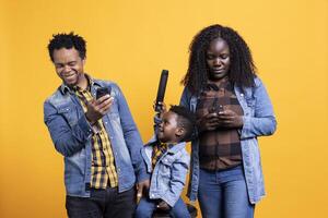 Happy proud parents using their smartphones next to their son in studio, cute toddler playing with a remote control. African american family having fun posing with multiple objects on camera. photo
