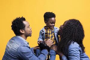 Happy small toddler playing around with a controller and sitting next to his parents, posing together in front of the camera. African american cheerful family of three against yellow background. photo
