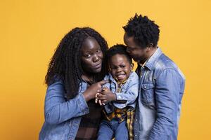 African american family and their little child in studio, holding each other against yellow background. Smiling proud young couple posing with their adorable toddler, enjoying leisure time. photo
