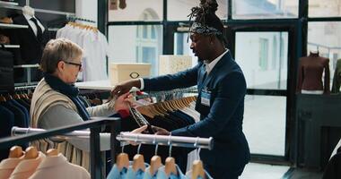 Elderly client asking to see multiple trendy bags in clothing store, talking to shop assistant about recommendations. African american worker shows different designer accessories. Camera B. photo