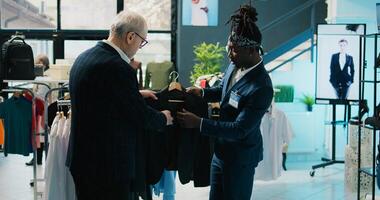 African american store assistant helping regular client with a suit in shopping center, senior customer looking to buy formal clothes. Elderly man shopping for new collection at the mall. Camera B. photo