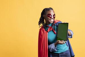 Portrait of smiling person portraying enigmatic superhero wearing mask, showcasing green screen tablet, studio background. Cheerful girl posing as hero with secret identity showing chroma key device photo