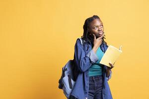 Portrait of BIPOC woman daydreaming while holding book, thinking of characters. Person with novel in arms caught in imagination while enjoying leisure time, isolated over studio background photo