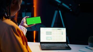 African american girl holds phone with isolated greenscreen at home, studying economics and finances at university. Young woman uses blank mockup template while she works on spreadsheets. Camera B. photo