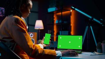 African american girl uses smartphone and pc with greenscreen, looking at modern devices running the same isolated mockup template on display. Young woman checks copyspace screens. Camera B. photo