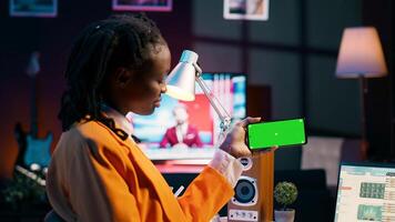 African american girl holds phone with isolated greenscreen at home, studying economics and finances at university. Young woman uses blank mockup template while she works on spreadsheets. Camera A. photo