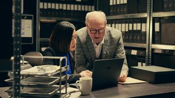Senior inspector showing case details to his partner in archive room, studying forensic evidence and classified criminal records in a box. Detectives team work on federal investigation. Camera B. photo