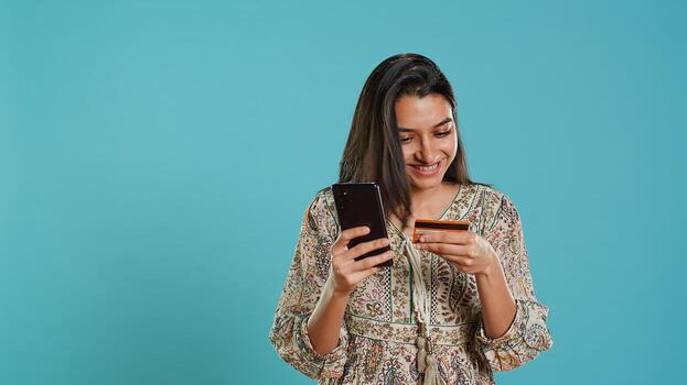 Indian woman excited to purchasing stuff, adding payment method on mobile phone website, isolated over studio background. Ecstatic client does internet shopping, typing debit card info, camera A photo