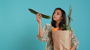 Woman with paper bag with vegetables testing quality, looking at cucumber, isolated over studio background. Person inspecting groceries after buying them from zero waste shop, camera A photo