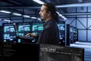 Engineer in data center using laptop to find firewall misconfigurations affecting rackmounts performance. Worker in server farm checking systems bottlenecks leading to sluggish data transfer rates photo