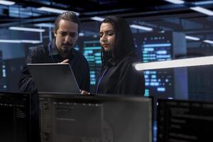 Programmers in server room checking recovery plan, monitoring tech gear energy consumption using notebook. Colleagues in data center workplace doing routine disk checks using laptop and tablet photo