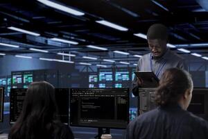 Multiethnic team in server room checking recovery plan and monitoring energy consumption. Diverse group of employees in data center location doing routine disk checks across components photo