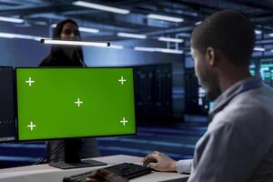 Technician using green screen desktop PC to oversee servers used for managing databases. African american man working on mockup computer monitor in data center storing information photo