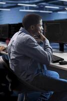 Man working on computer in modern data center, sitting at desk and thinking of solutions. Employee doing IT infrastructure management on server racks in high tech environment using PC photo