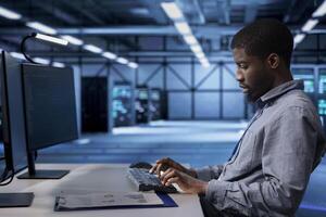 Employee checking for signs of imminent tech failure in data center supercomputers. African american man working at PC desk to prevent damages in server farm, monitoring critical systems photo