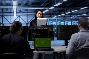Manager inspecting server room equipment, reading paperwork from folder while engineers work on isolated screen laptop. Supervisor checking data center files next to chroma key device on desk photo