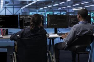 Person with paraplegia and colleague working in inclusive server hub workspace, typing on PC. Woman in wheelchair and coworker doing tasks in data center housing advanced data storage infrastructure photo
