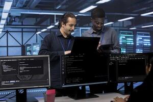 Coworkers in data center using laptop and tablet to find firewall misconfigurations affecting servers. Colleagues in server room checking systems bottlenecks leading to sluggish data transfer rates photo