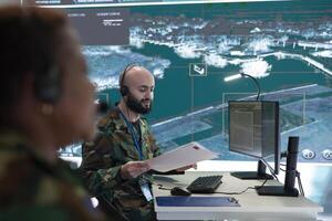 Skilled IT engineer monitoring satellite CCTV footage recorded on computer and big screen in military command post. Army trooper working in the reconnaissance division for information. photo
