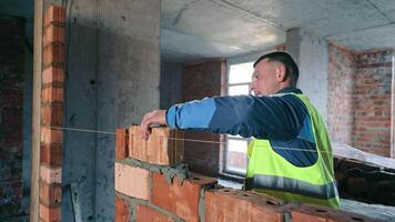 A mason in a vest meticulously building a brick wall inside a construction site. The builder demonstrates the skill and precision required for masonry. video