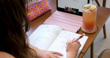 conectados Aprendendo menina dentro uma cafeteria trabalhando em uma Rosa tábua escrevendo baixa matemático equações com uma caneta bebendo uma coquetel dentro uma tropical país relaxante enquanto estudando estude conectados video
