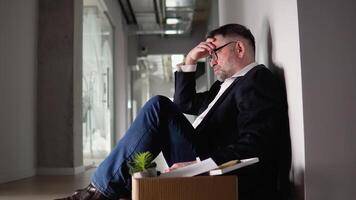 Fired senior employee sitting frustrated and upset in the hallway near office with dismissal box and drinks alcohol. He lost work or retiring video