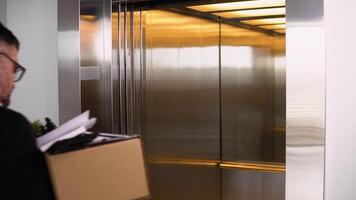 Sad dismissed senior worker with carton dismissal box getting into the elevator. Old man is retiring. Retirement concept. Dismissal from work video
