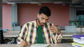 Preparing to the exams. Thoughtful tired young indian student writing something in her note pad and reading book while sitting at the library desk video
