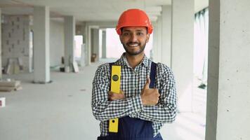 Portrait of Indian worker builder in uniform wearing a safety helmet with waterpas looking at the camera, standing on construction site of office center video