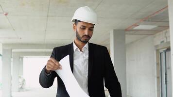 Serious attractive indian industrial engineer in hard hat with project drawings at construction of a skyscraper. Professional and industry, builder architect job, worker checking work at plant indoor video