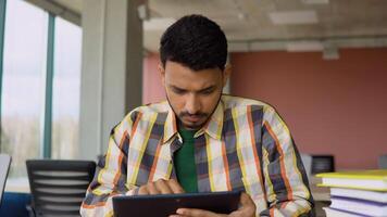 Indian student is studying in a college library. He uses a tablet and studies online video