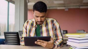 Indian student is studying in a school library. He uses a tablet and studies online video