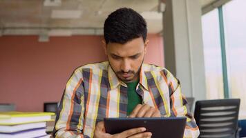 Indian student is studying in a university library. He uses a tablet and studies online video