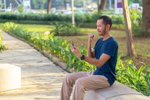 Young asian men with blue shirt smiling and laughing while playing smartphone on the park outdoor photo