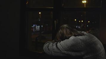 Lonely young woman looks out night window. Media. Young woman sits on window and looks sadly at night city. Woman with depression sits alone on window of house photo
