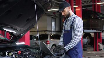 Experienced mechanic in uniform is using a laptop while repairing car in auto service video