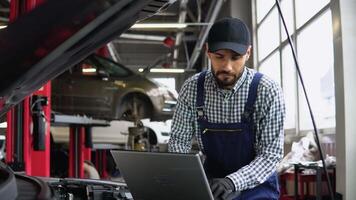 Experienced mechanic in uniform is using a laptop while repairing car in auto service video