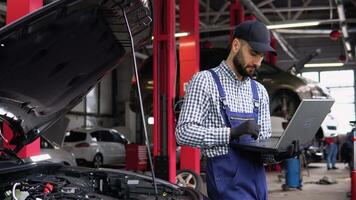 Experienced mechanic in uniform is using a laptop while repairing car in auto service video