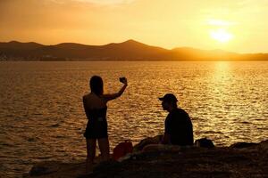 Silhouette of a man and a woman taking a selfie on the beach at sunset . Group of happy Asian friends woman and man using smartphone to take a photo and together on beach with sunset during summer