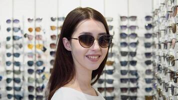 Portrait of young woman in new sunglasses in optics on a background of showcase with different models of glasses video