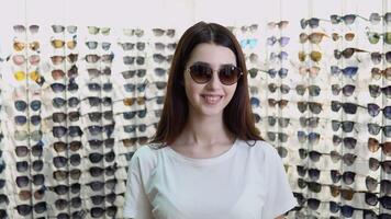 Portrait of young woman in new sunglasses in optics on a background of showcase with different models of glasses video