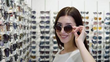 Young woman trying on new sunglasses in optical store on a background of showcase with different models of glasses video