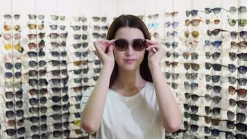 Young woman trying on new sunglasses in optical store on a background of showcase with different models of glasses video