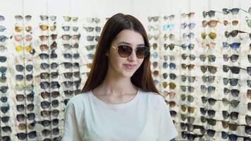 Portrait of young woman in new sunglasses in optics on a background of showcase with different models of glasses video