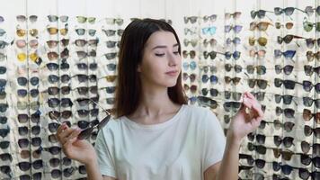 Portrait of cheerful hesitating woman in optician store, making decision, holding stylish sunglasses, choosing what she should buy. Girl on a background of showcase with different models of glasses video