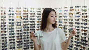 Portrait of cheerful hesitating woman in optician store, making decision, holding stylish sunglasses, choosing what she should buy. Girl on a background of showcase with different models of glasses video