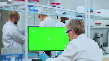 Elderly researcher looking at chroma key display in modern equipped lab. Multiethnic team of microbiologists doing vaccine research writing on device with green screen, isolated, mockup display. video