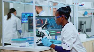 Black biochemist woman sitting in modern equipped lab analysing blood tests. Multiethnic team examining virus evolution using high tech for scientific research of treatment development against covid19 video